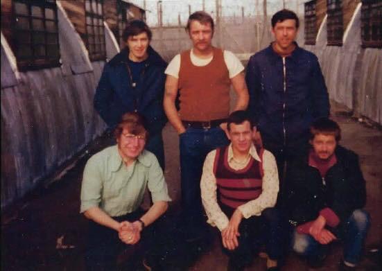 A group of six men posing together, with three seated in the front and three standing behind them, in a rustic setting.