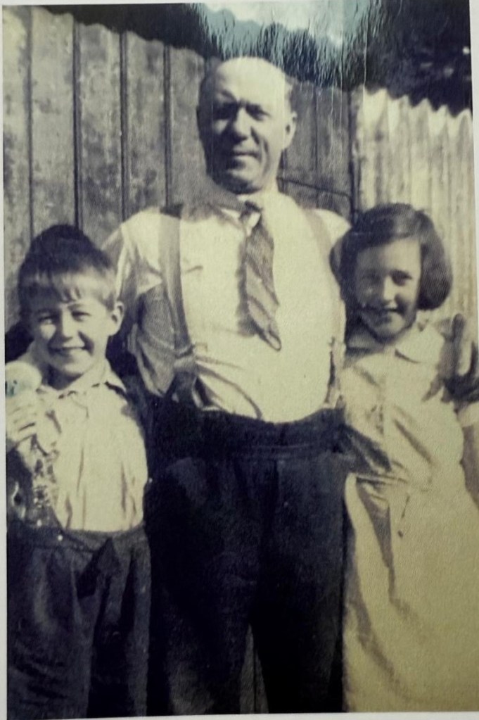 A black and white photo of a man standing in the middle with two children, a boy on the left and a girl on the right, all smiling. The man is wearing a white shirt, tie, and suspenders, while the children are dressed in light-colored outfits.