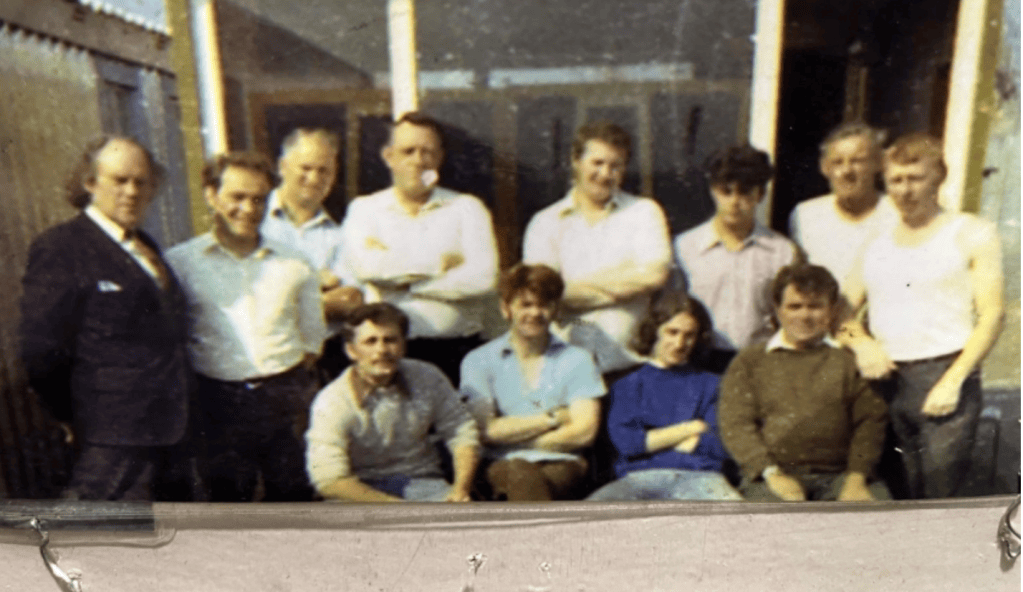 A group photo of thirteen people posing for the camera outdoors, with some standing and others sitting. They appear to be men and women dressed casually, with a mixture of smiles and serious expressions, in front of a building.