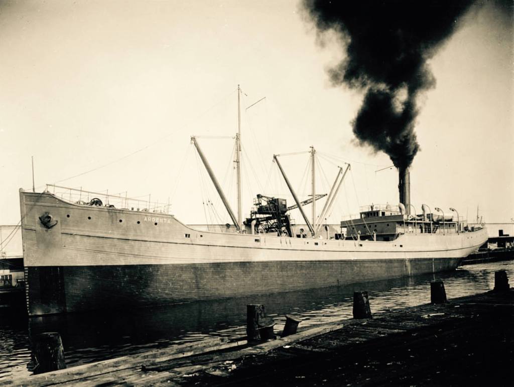 Black and white image of a large steamship, with smoke billowing from its smokestack, docked at a harbor.