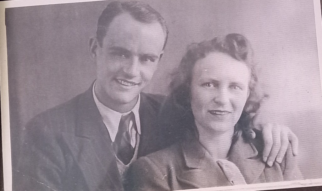 Black and white photo of a smiling couple posing closely together. The man is wearing a suit with a tie, and the woman has curly hair and is dressed in formal attire.